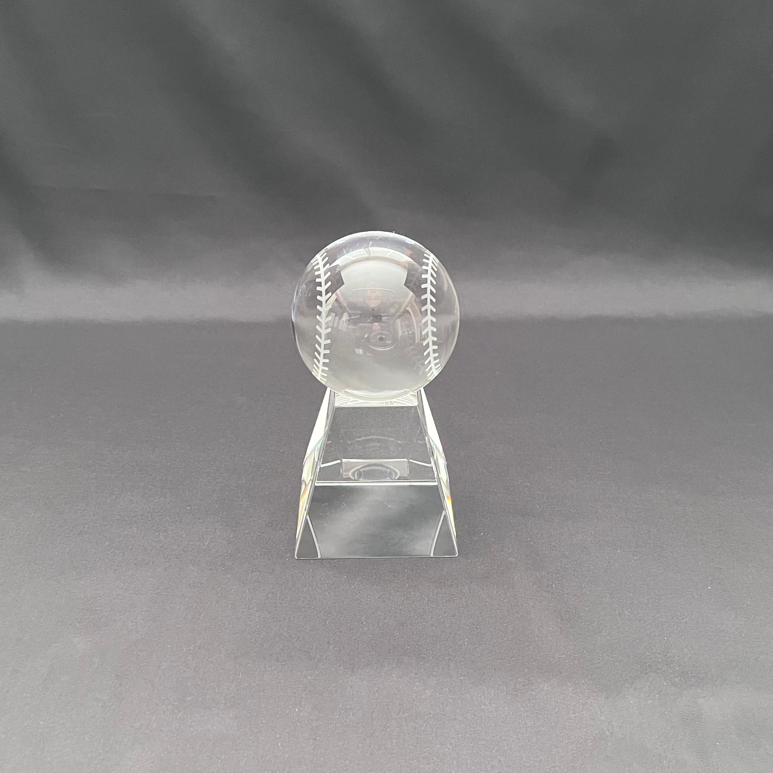 A clear crystal baseball trophy on a matching clear acrylic stand, photographed against a dark gray fabric backdrop.