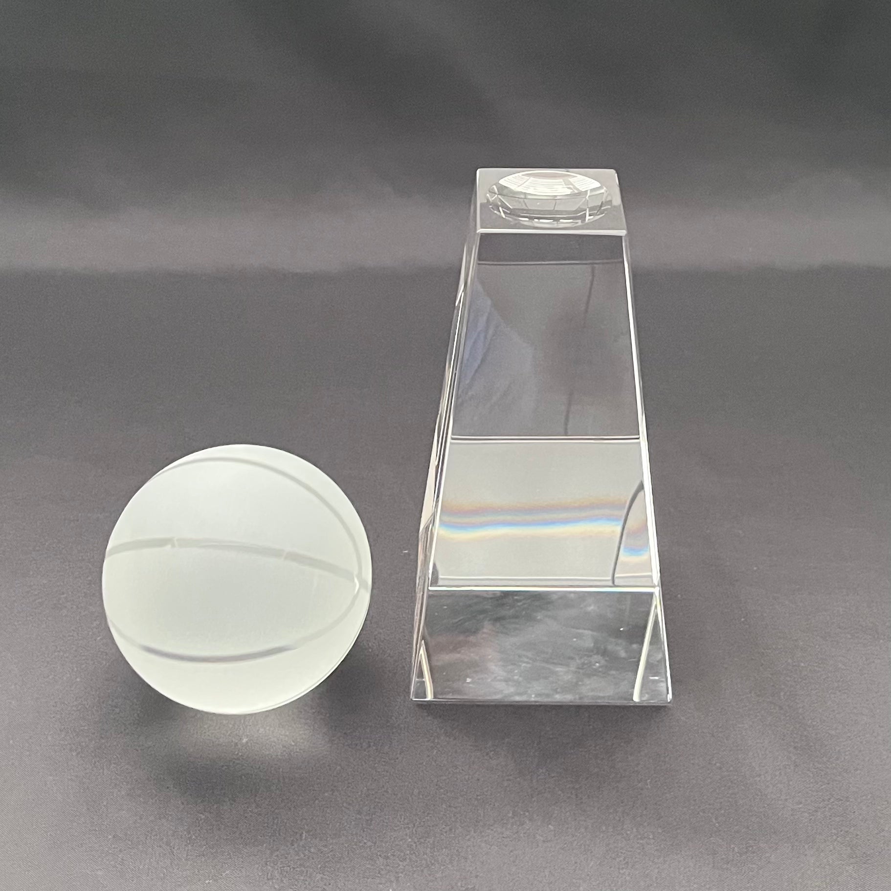 A clear acrylic or glass decorative object set on a dark gray background, featuring a round frosted ball with horizontal grooves and a tall rectangular prism with a circular inset at the top.