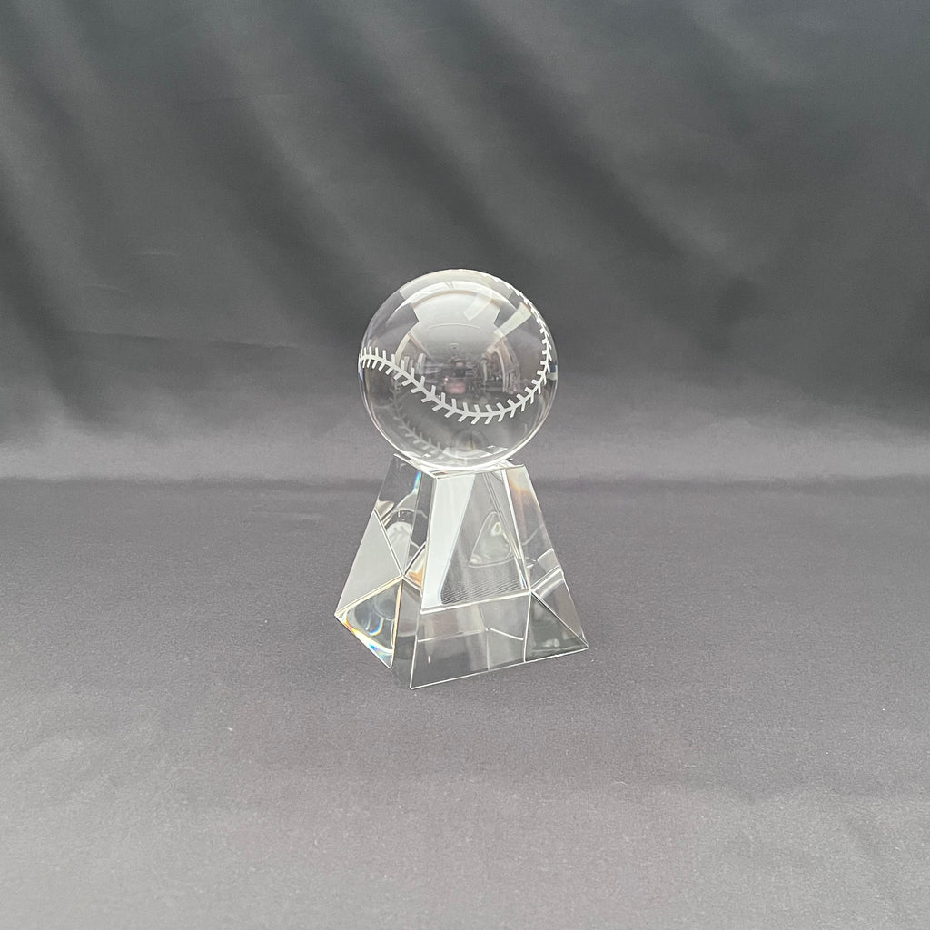 A clear crystal baseball trophy stand featuring a transparent baseball with white stitching, set against a dark gray fabric backdrop.