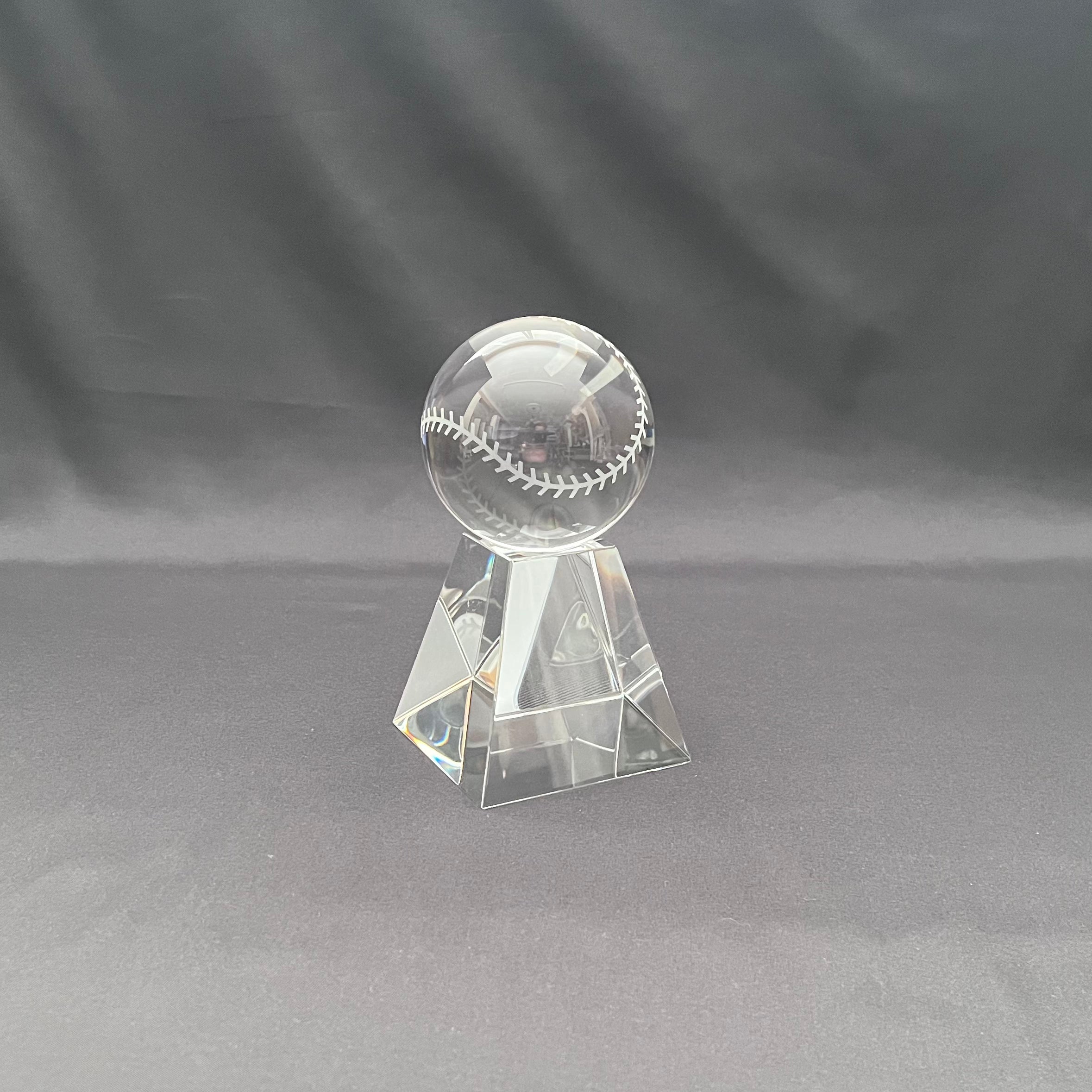 A clear crystal baseball trophy stand featuring a transparent baseball with white stitching, set against a dark gray fabric backdrop.
