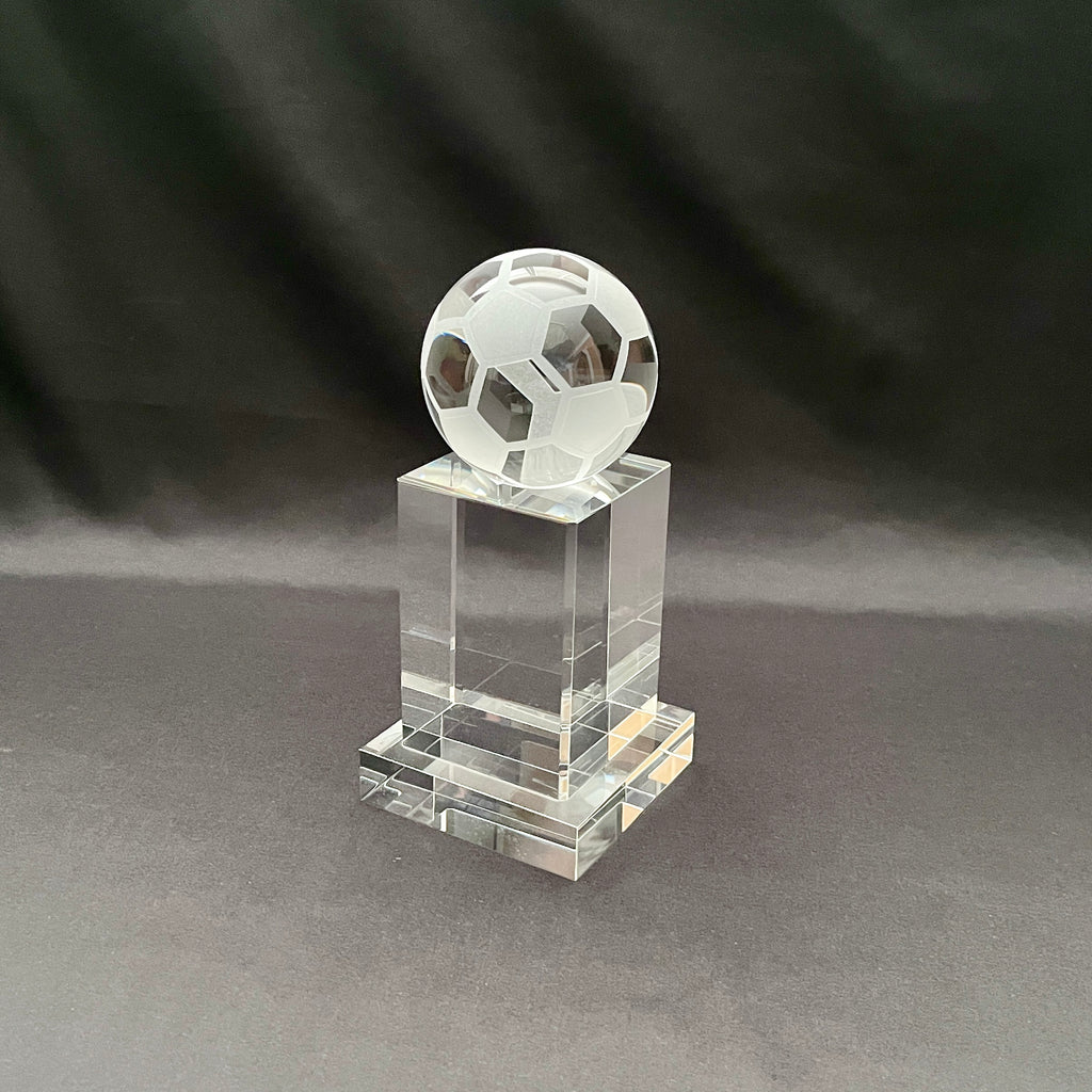 A clear crystal trophy stand featuring a faceted soccer ball sculpture on top, set against a dark gray fabric backdrop.