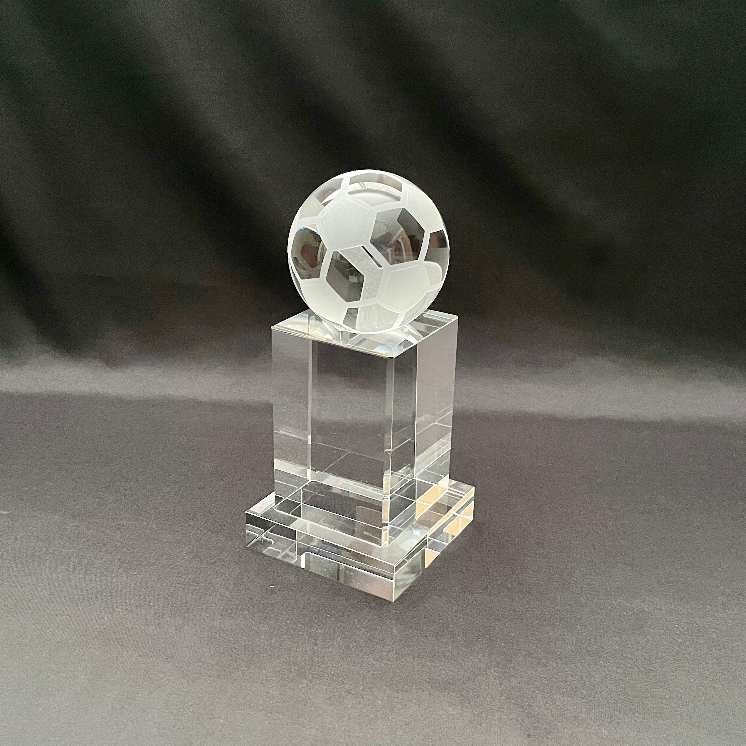 A clear crystal trophy stand featuring a faceted soccer ball sculpture on top, set against a dark gray fabric backdrop.