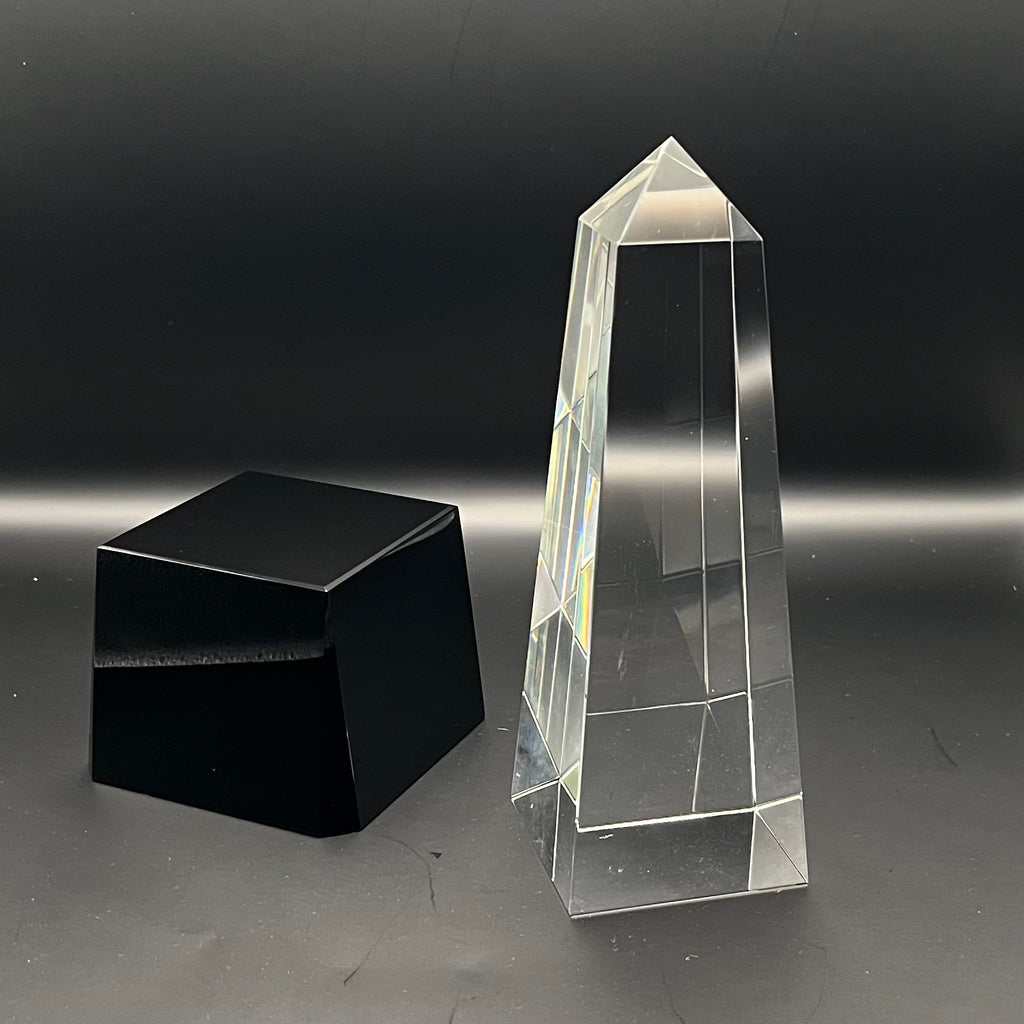 Clear crystal obelisk and black cube on a reflective surface with a dark background