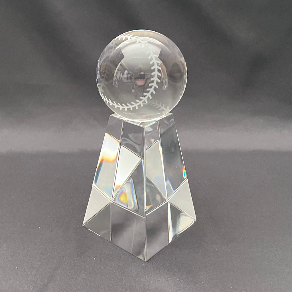 A clear glass baseball trophy stand with a faceted geometric base and a transparent round top engraved with a baseball design, set against a dark gray background.