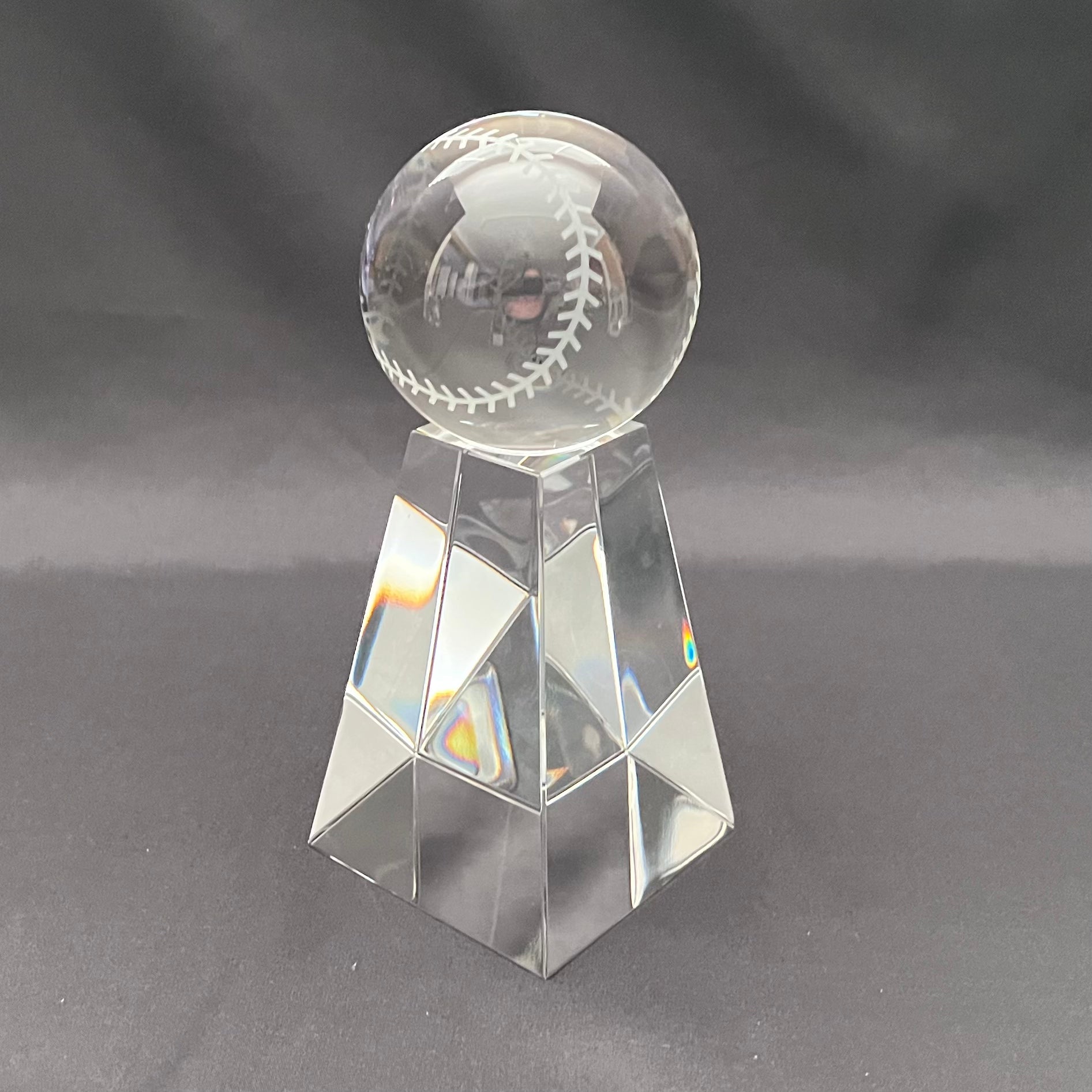A clear glass baseball trophy stand with a faceted geometric base and a transparent round top engraved with a baseball design, set against a dark gray background.