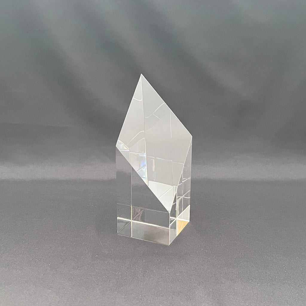 A clear acrylic or glass geometric crystal award block with a faceted diamond-like top, standing on a gray fabric background.